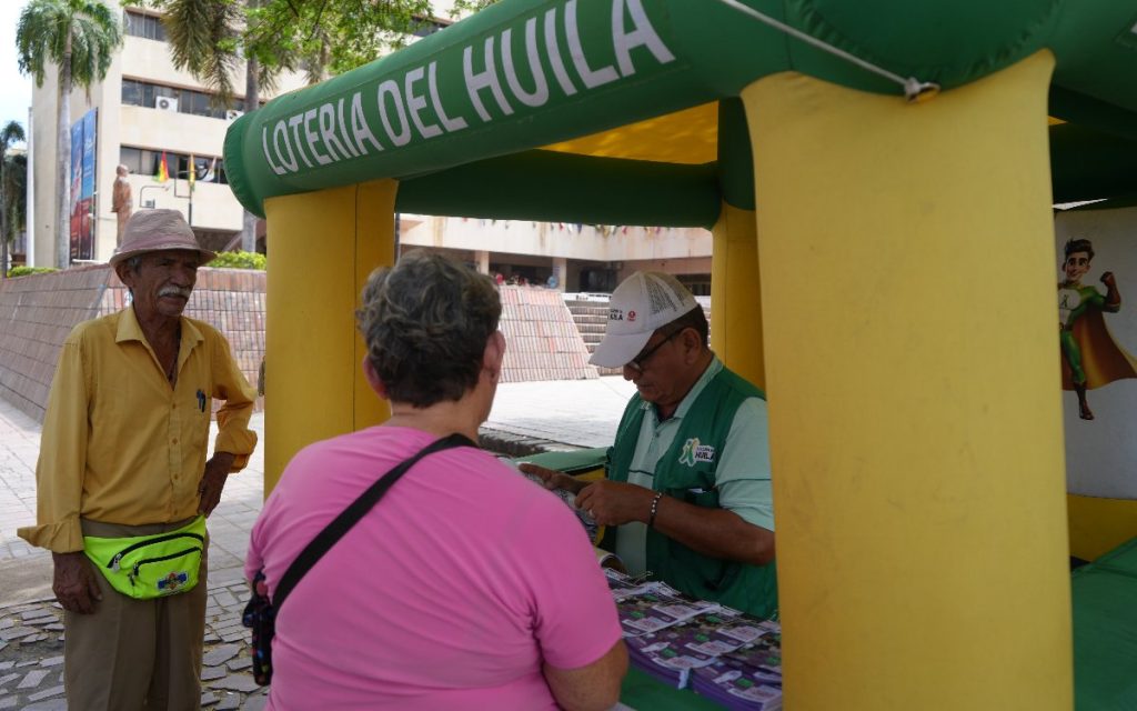Fotografía en exterior que muestra un estand inflable de colores verde y amarillo con la inscripción "Lotería del Huila". Dentro del estand, un vendedor con gorra y chaleco institucional organiza los billetes mientras atiende a una mujer vestida de rosa en primer plano y a un hombre mayor con sombrero y camisa amarilla de pie al lado del estand.
