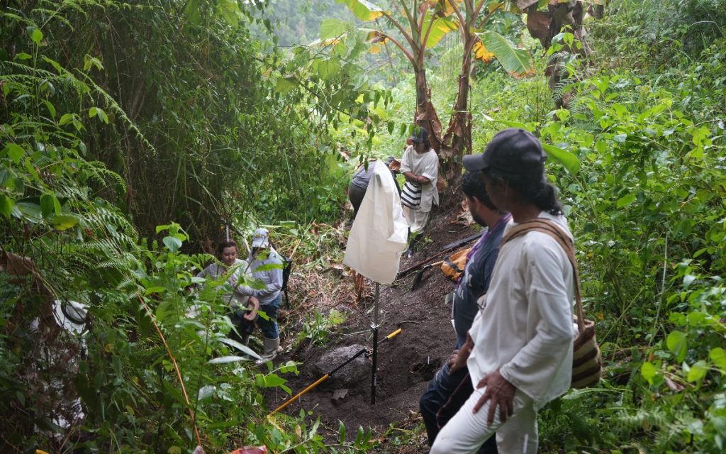 Vista panorámica en pendiente rodeada de frondosa vegetación. Varios técnicos y miembros de la comunidad observan los trabajos en una zanja excavada en el suelo, la cual cuenta con un soporte e instrumento técnico cubierto con un protector claro.