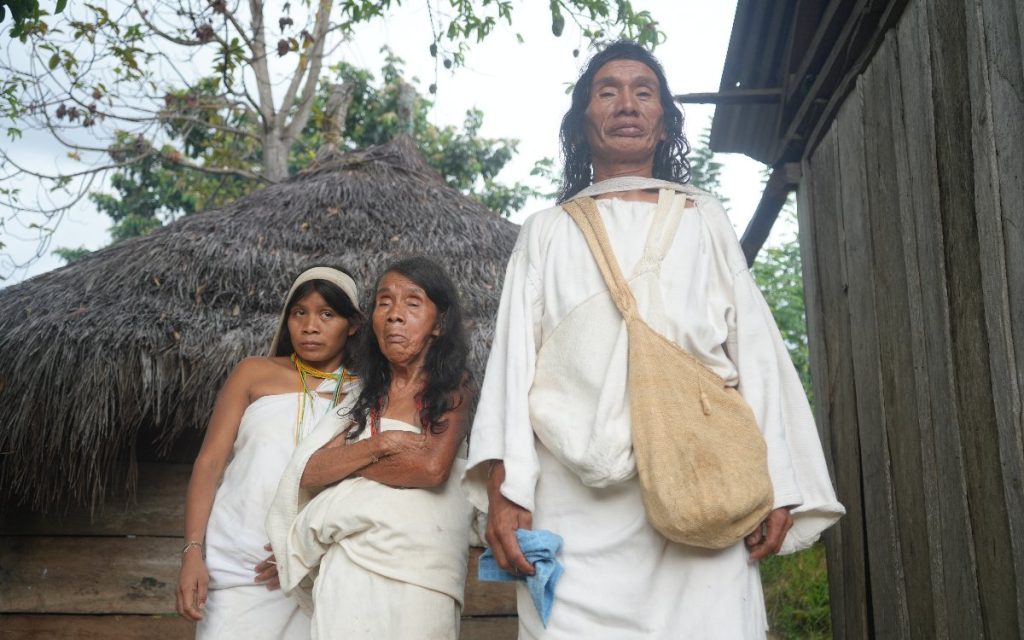 Retrato de tres personas indígenas frente a su vivienda tradicional en la Sierra Nevada. En primer plano destaca un hombre alto vistiendo la manta blanca tradicional y una mochila cruzada sobre el pecho; a su lado se encuentran dos mujeres con vestidos blancos tradicionales e indumentaria autóctona.