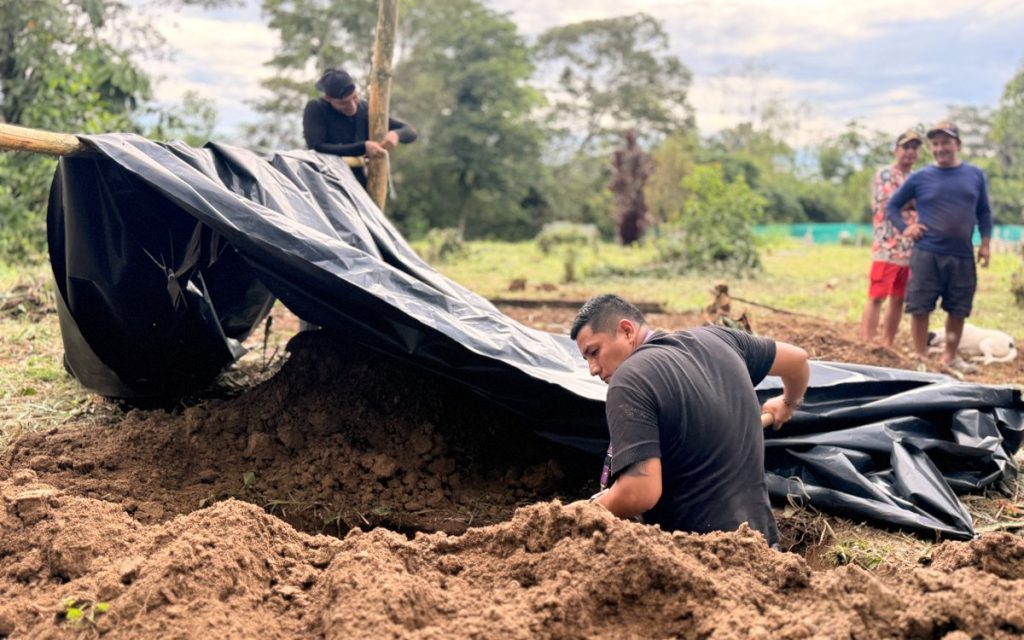 Vista en primer plano de un excavador agachado junto a un montón de tierra extraída, acomodando una gran lona plástica negra sostenida por varas de madera. En el fondo, dos hombres observan la labor en un terreno de campo rodeado de árboles.