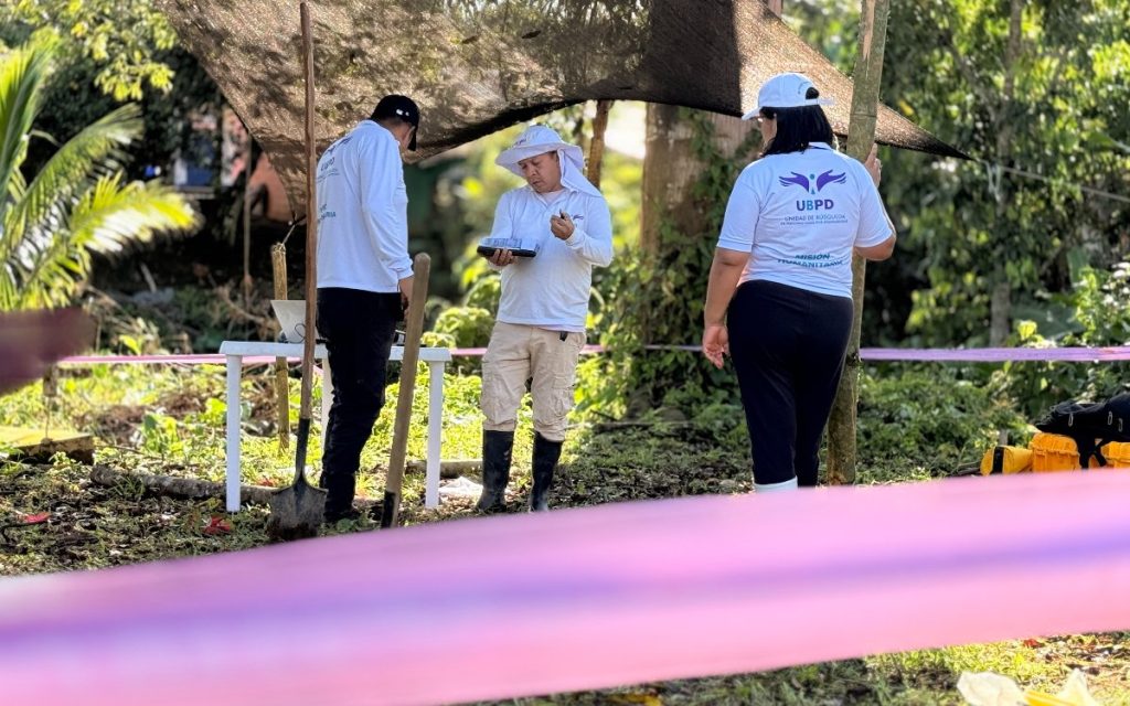 Tres personas con camisetas blancas identificadas con el logo de la UBPD y el texto "Misión Humanitaria" trabajan bajo la sombra de un toldo. Una persona revisa un cuaderno o libreta, mientras las otras dos observan el área delimitada con una cinta.