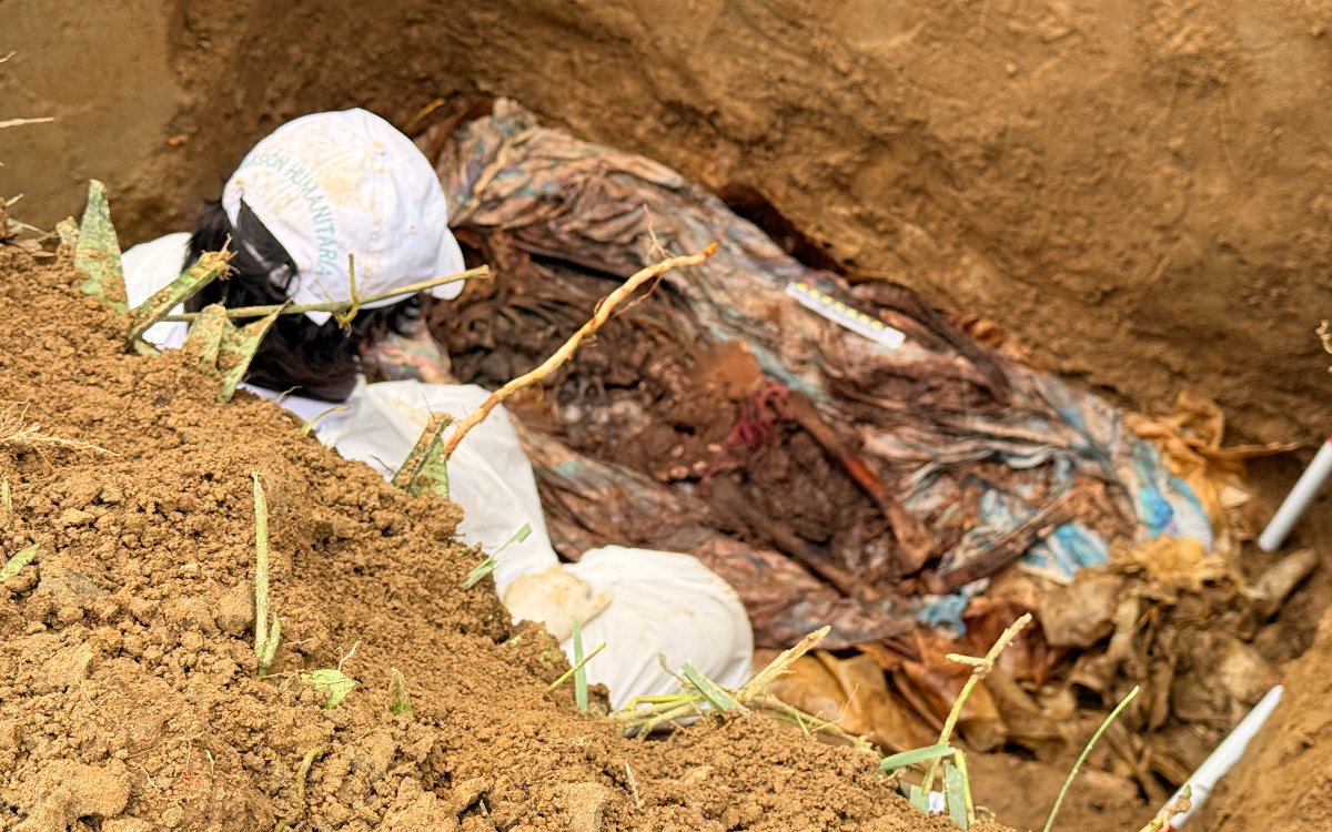 Toma aérea de un profesional forense vestido con traje y gorra blanca de la misión humanitaria, agachado dentro de una excavación en la tierra para examinar cuidadosamente los restos y evidencias halladas en el suelo.