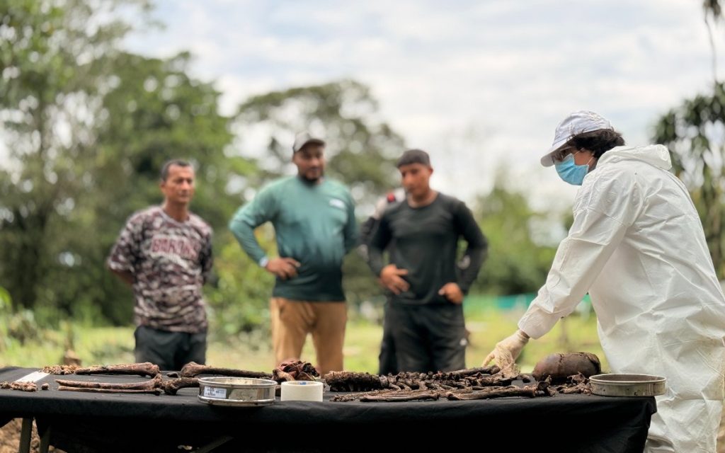 En primer plano, un experto forense con traje biológico blanco, tapabocas y guantes dispone y analiza restos óseos sobre una mesa cubierta con tela negra. En segundo plano, tres hombres vestidos de civil observan con atención el procedimiento al aire libre.