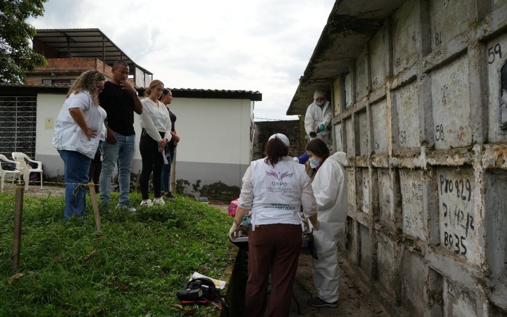 A la izquierda, un grupo de familiares y una funcionaria de la UBPD observan atentamente el trabajo de exhumación. A la derecha, forenses con trajes de bioseguridad manipulan hallazgos en una mesa dispuesta frente a la pared de bóvedas numeradas.