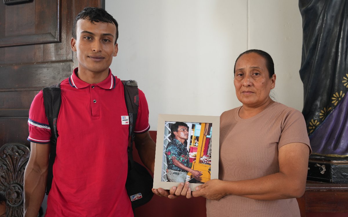 Fotografía de interior en la que un joven a la izquierda (con camiseta polo roja y morral) y una mujer a la derecha (con camiseta beige) sostienen en el centro un marco con la foto en color de Cruz Arcángel Villada. Al fondo se aprecia una puerta de madera y una escultura religiosa.