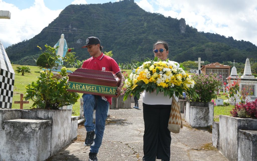 Imagen al aire libre en un cementerio rural. Un joven con gorra negra y camiseta roja camina cargando un pequeño cofre fúnebre de madera con la inscripción "Cruz Arcángel Villada". A su lado, una mujer con gafas de sol lleva un gran ramo de flores amarillas y blancas. Al fondo se observa una estatua de la Virgen María y una montaña bajo un cielo despejado.