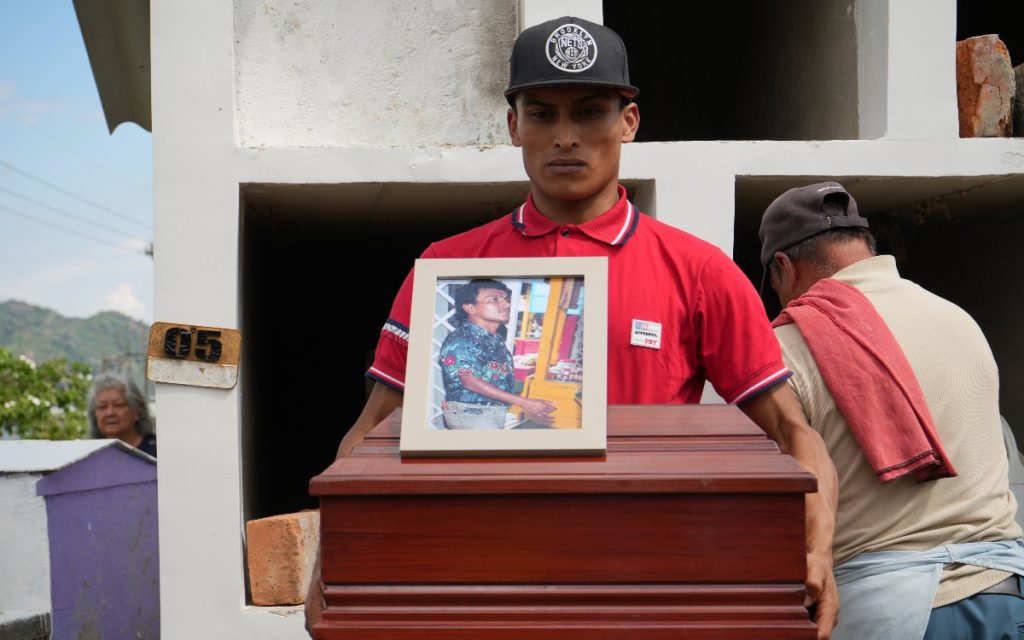 Plano medio de un joven con gorra negra y camiseta roja sosteniendo el pequeño cofre fúnebre de madera. Sobre la parte superior del cofre descansa el retrato enmarcado del hombre desaparecido. A la derecha, un operario trabaja de espaldas en la apertura de una bóveda en el cementerio.