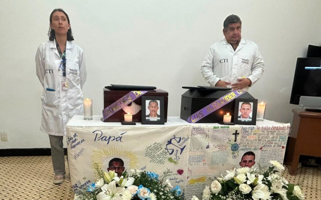 Dos funcionarios del CTI de la Fiscalía, vestidos con batas blancas de laboratorio, permanecen de pie tras una mesa cubierta con un mantel blanco repleto de mensajes y dibujos hechos a mano. Sobre la mesa reposan dos urnas de madera, cada una con una cinta púrpura, una fotografía de la persona fallecida y una veladora encendida. En la base del altar hay arreglos florales blancos y azules.