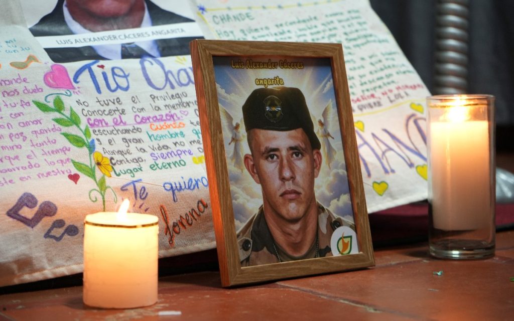 Fotografía enmarcada en madera de Luis Alexander Cáceres Angarita usando gorra y ropa militar. A los lados hay dos veladoras cilíndricas encendidas. Detrás del marco se aprecia una manta o cartel con textos manuscritos con dedicatorias de sus familiares, como "Tío Chande" y "Te quiero".