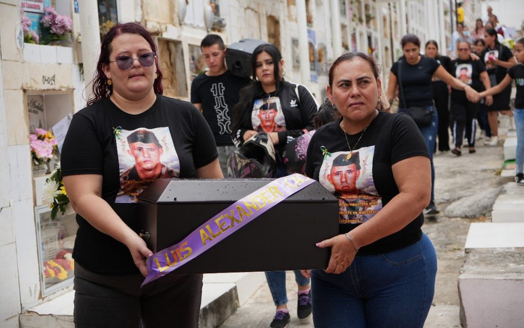 Dos mujeres avanzan por el pasillo de un cementerio cargando juntas una urna funeraria de madera con una cinta morada que dice "LUIS ALEXANDER CÁCERES". Ambas visten camisetas negras personalizadas con la fotografía de la víctima y el mensaje "permanecerá vivo en nuestros corazones". Detrás de ellas caminan más familiares acompañando el cortejo.