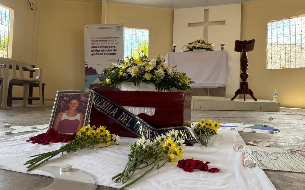 En primer plano, sobre una manta blanca en el suelo de una capilla, reposa un cofre de madera para cenizas o restos, adornado con una cinta azul grabada con el nombre "Luzmila del Carmen Atencia". A su lado hay un retrato enmarcado de una mujer, ramos de crisantemos amarillos y blancos, y pétalos de rosas rojas. Al fondo se observa el altar con una cruz blanca y un pendón institucional de búsqueda de personas.