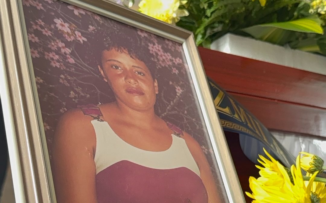 Plano detalle de una fotografía enmarcada que muestra el retrato de una mujer con cabello corto y tez morena, vestida con una prenda de tirantes en tonos blanco y borgoña. El marco se encuentra rodeado por crisantemos amarillos y flores en el fondo.
