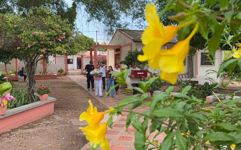 Vista de un grupo de hombres y mujeres caminando por un camino empedrado al aire libre, rodeado de vegetación y construcciones blancas de teja. Una de las personas lleva en sus manos el cofre fúnebre de madera, mientras que otras cargan coronas y ramos de flores. En primer plano resaltan flores amarillas desenfocadas de un arbusto.