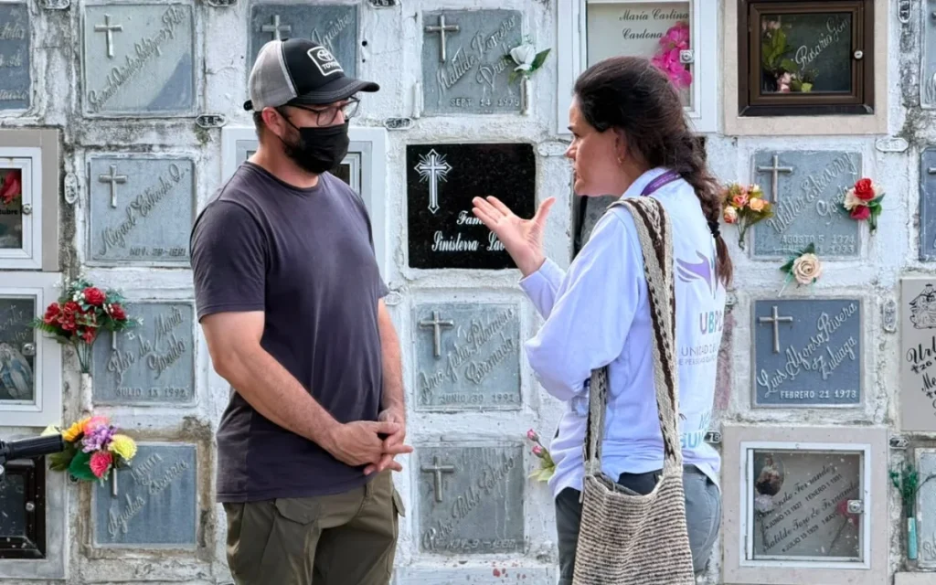 Un hombre con camiseta oscura, gorra de béisbol y tapabocas negro escucha atentamente a una funcionaria de la Unidad de Búsqueda de Personas dadas por Desaparecidas (UBPD). Ella gesticula con las manos mientras dialogan en un cementerio, teniendo como fondo varios nichos funerarios con lápidas y flores.