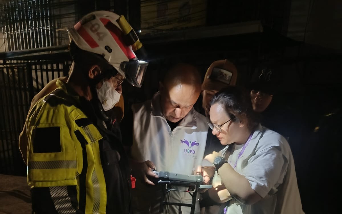 Fotografía nocturna en la que un rescatista equipado con casco de protección y chaqueta reflectante observa detenidamente la pantalla de un dispositivo de control junto a dos funcionarios de la UBPD y otros colaboradores.