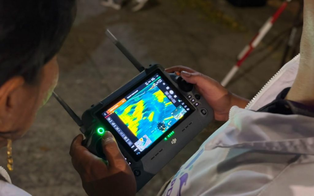 Toma en ángulo picado sobre los hombros de la operadora, donde se aprecia la pantalla digital del control del dron transmitiendo una imagen termográfica con variaciones de temperatura representadas en tonos cálidos y fríos.