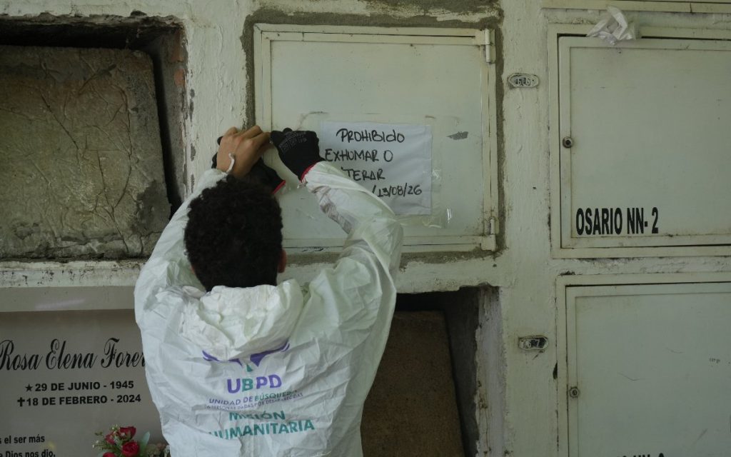 Vista posterior de un funcionario con traje blanco con el logotipo "UBPD Misión Humanitaria", pegando un letrero impreso que dice "PROHIBIDO EXHUMAR O ALTERAR" sobre la tapa de una bóveda en el cementerio.