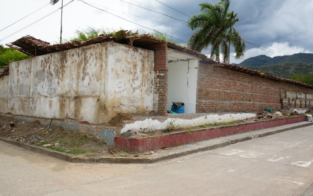 Fachada exterior de una edificación de ladrillo y concreto en una esquina de calle pavimentada. La estructura muestra daños significativos, con partes del techo de teja destruidas y agujeros en los muros de contención.