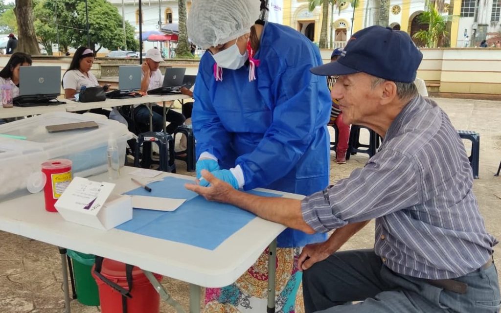 En un punto de atención al aire libre, un profesional de la salud con bata quirúrgica azul, tapabocas, gorro y guantes sostiene la mano de un hombre mayor para tomarle una muestra de sangre capilar en el dedo. Sobre la mesa blanca hay un contenedor rojo para desechos biológicos, cajas de insumos médicos y equipos de cómputo.
