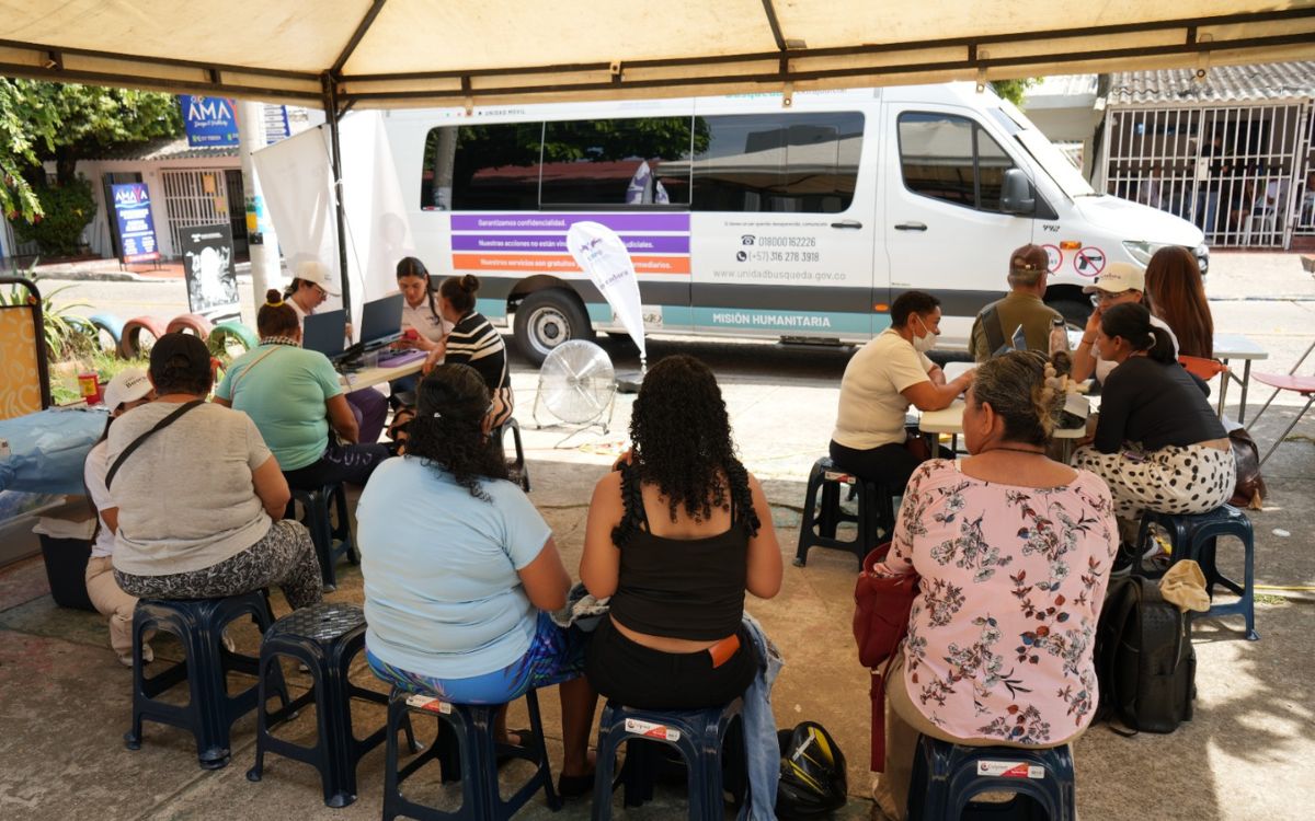 Jornada de atención comunitaria bajo una carpa, donde personas sentadas en bancos plásticos reciben orientación de funcionarios frente a computadores portátiles, junto a una camioneta tipo van rotulada con la frase Misión Humanitaria.