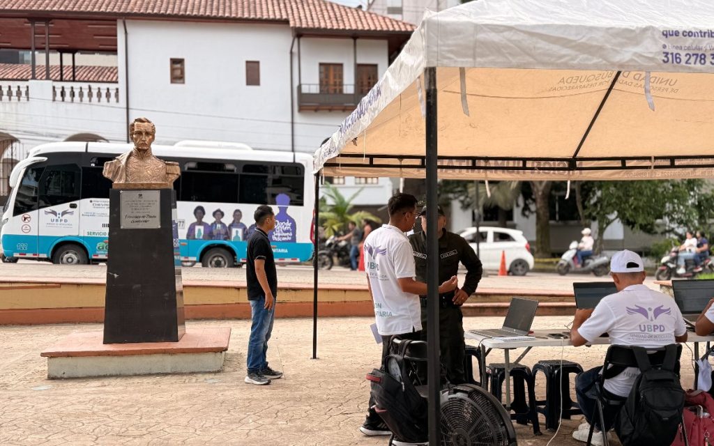 Plaza pública donde se observa un busto en bronce sobre un pedestal dedicado a Francisco de Paula Santander. A la derecha hay una carpa con personal atendiendo con computadores portátiles y conversando con un agente de policía. Al fondo se ve un autobús de la UBPD y una edificación colonial de paredes blancas con balcones de madera.
