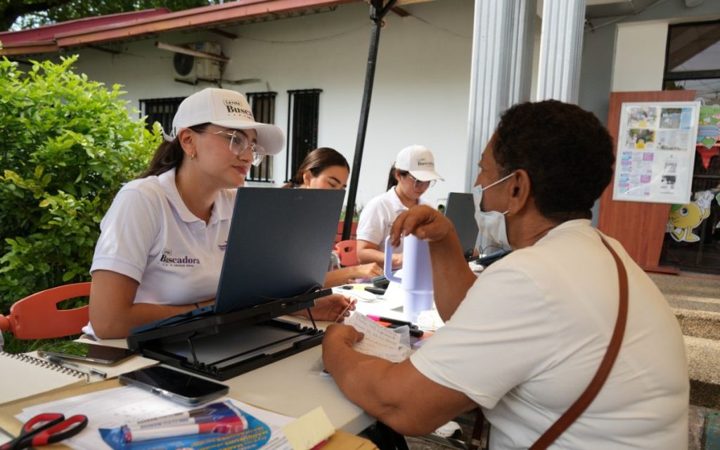 Funcionaria con polo y gorra blanca de La Ruta Buscadora atiende y escucha a una ciudadana frente a su computador portátil en una mesa al aire libre.