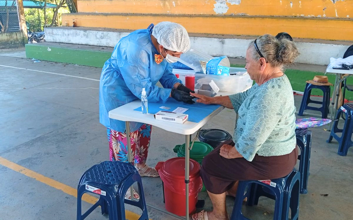 Personal de la Unidad de Búsqueda con indumentaria realiza una toma de muestra de sangre en el dedo a una mujer adulta mayor durante una jornada médica.