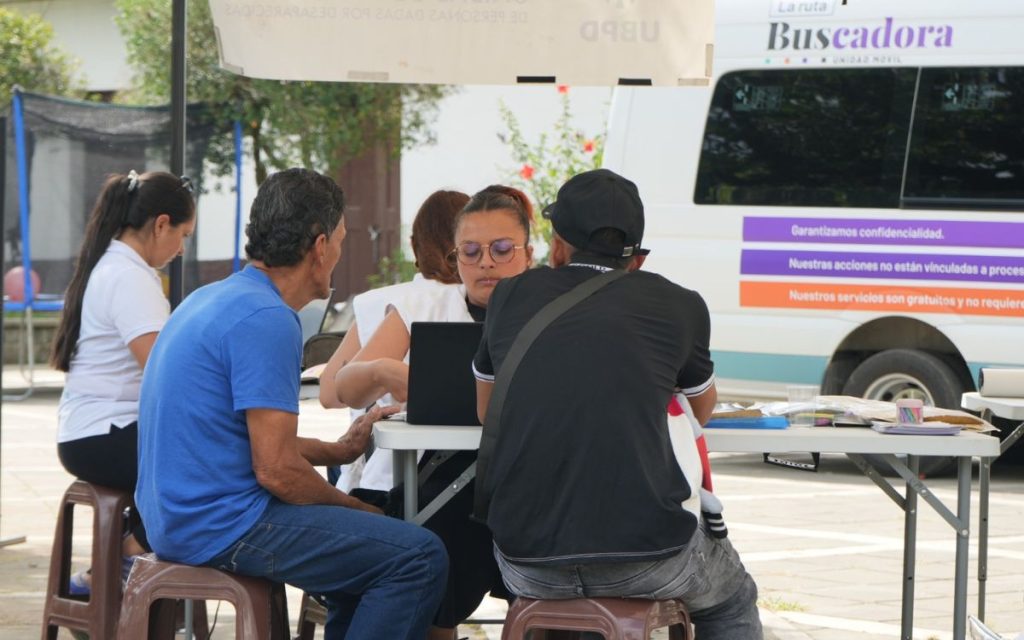 Una funcionaria de la UBPD con gafas y un computador portátil atiende a dos personas sentadas frente a ella en una mesa de atención al aire libre, junto a la unidad móvil Ruta Buscadora.