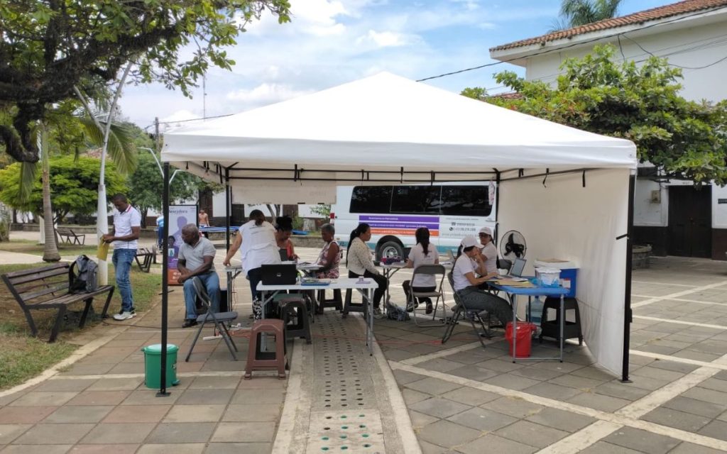 Carpa blanca instalada en una plaza pública con mesas y personal de la UBPD atendiendo a varias personas de la comunidad; al fondo se observa la unidad móvil de la Ruta Buscadora.