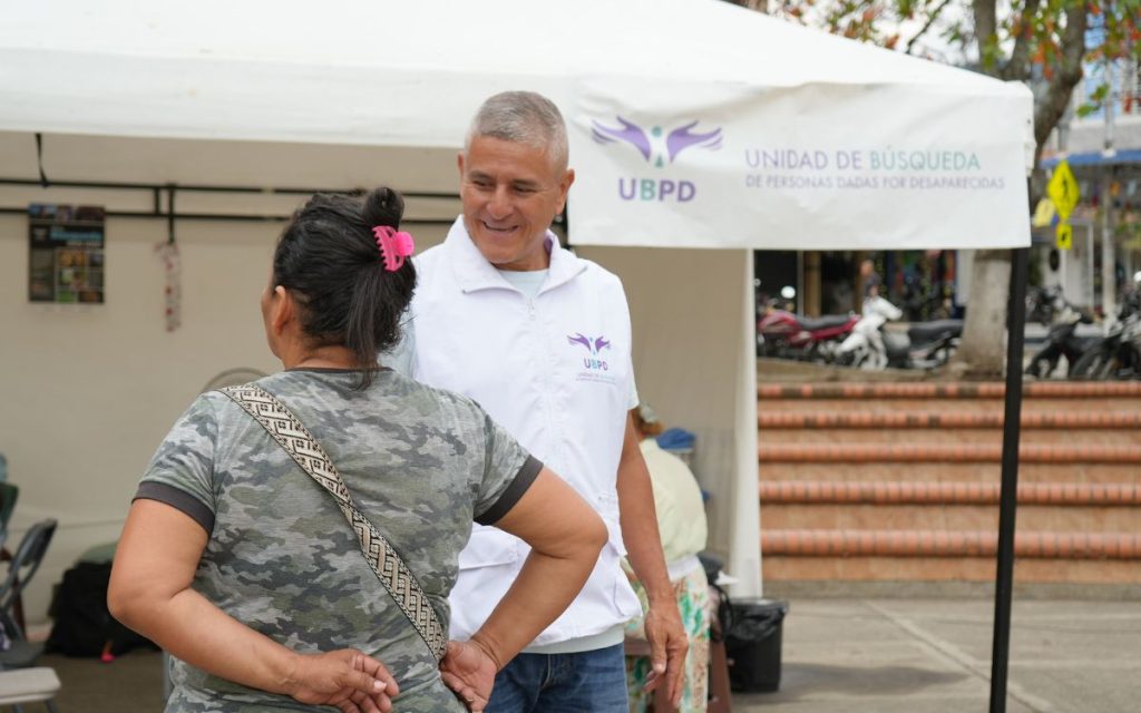 Un funcionario de la UBPD con chaleco blanco institucional sonríe mientras conversa frente a frente con una mujer en el punto de atención al aire libre.