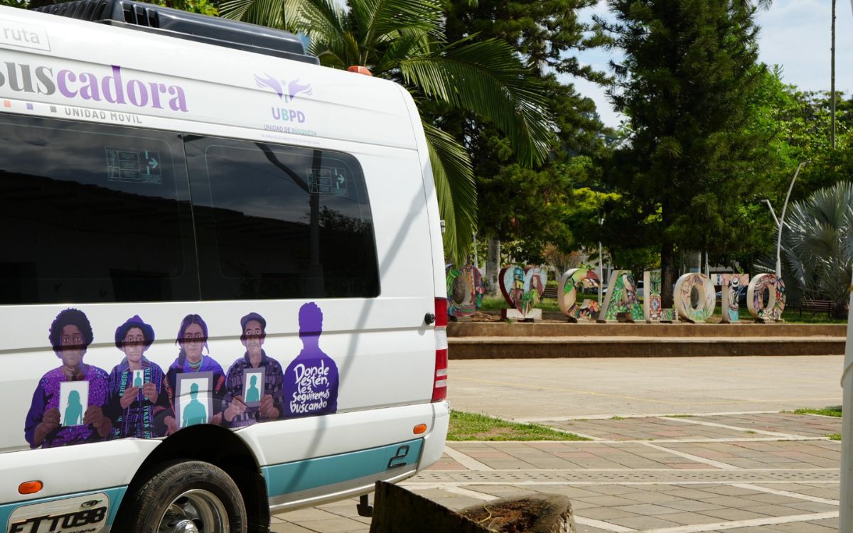 Costado trasero de la unidad móvil de la Ruta Buscadora de la UBPD, decorada con ilustraciones de personas sosteniendo retratos y la frase «Donde estén, les seguiremos buscando», estacionada frente al letrero turístico de Caloto en un parque arbolado.