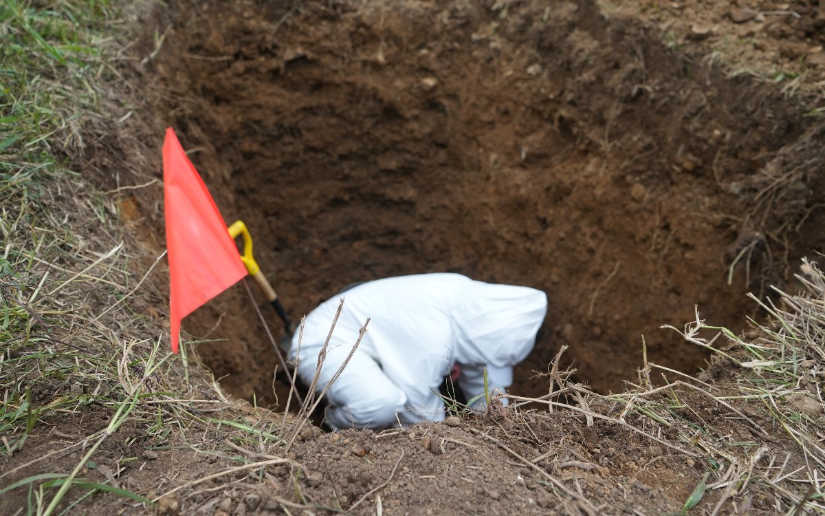 Toma superior de un foso o excavación en terreno de tierra y hierba. En el borde izquierdo hay una pequeña bandera roja con su mástil incrustado junto al mango de una pala. En el fondo de la excavación se observa a un especialista agachado realizando labores forenses o de búsqueda.