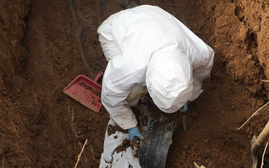 Primer plano cenital dentro de una excavación donde una persona especializada, totalmente cubierta con traje de bioseguridad blanco y guantes azules, manipulate tierra sobre una superficie blanca. Al lado se aprecian herramientas de trabajo de precisión, como un recogedor plástico rosa y un balde pequeño.