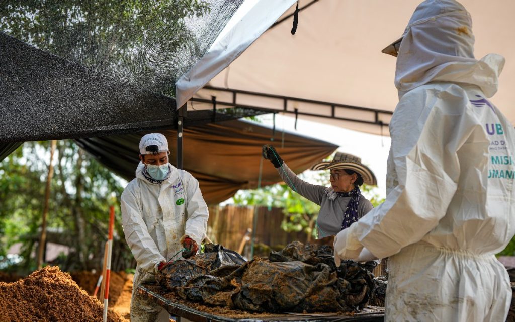 Dos expertos en forense usan trajes de protección biológica blancos y mascarillas bajo un toldo o carpa de campo. A la izquierda, un hombre revisa materiales envueltos sobre una mesa, mientras que en el centro una mujer con sombrero tradicional vueltiao observa minuciosamente el proceso. A la derecha, se aprecia la espalda de otra persona con el logo oficial de la UBPD y el texto "Misión Humanitaria".