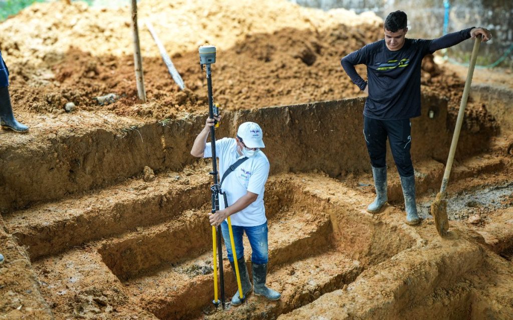 Un profesional capacitado de la UBPD, que viste camiseta blanca con distintivos de la entidad, gorra y botas de caucho, opera una herramienta GPS de precisión para georreferenciar una excavación en tierra. A su derecha, apoyado sobre una pala de mano en el borde de la fosa, un hombre con camiseta manga larga negra observa el procedimiento técnico.