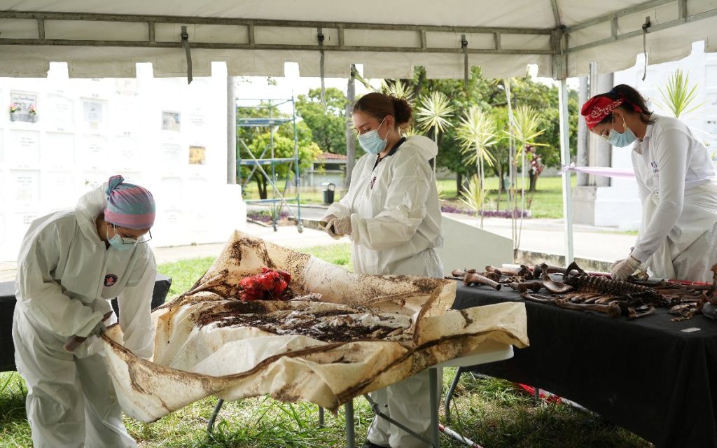 Tres profesionales con trajes de protección blancos y mascarillas trabajan bajo una carpa. Una de ellas manipula una sábana con sedimentos sobre una mesa plegable, mientras que otra organiza osamentas en una mesa contigua. De fondo se observa la infraestructura blanca de un cementerio y zonas verdes.