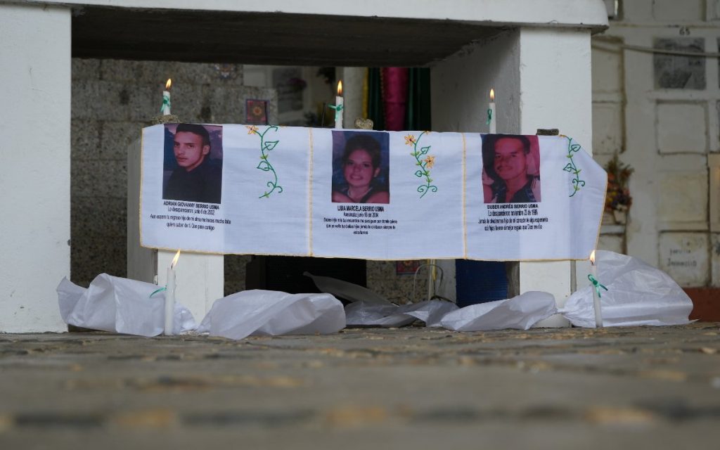 Un pequeño altar instalado al pie de una bóveda. Sobre un paño blanco con bordados florales se aprecian tres fotografías de rostros acompañadas de textos alusivos a sus búsquedas: "Adrian Giovanny Berrío Usma", "Libia Marcela Berrío Usma" y "Duber Andrés Berrío Usma". Flanqueando el altar hay cuatro velas encendidas colocada sobre el suelo de piedra.