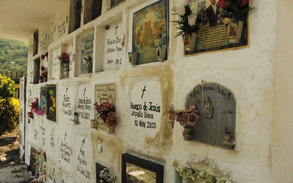Sección de un cementerio con bóvedas decoradas de forma personalizada. Los nichos blancos contienen fotografías enmarcadas de los difuntos, pequeñas esculturas e imágenes religiosas, placas conmemorativas y flores artificiales. En el centro destaca la inscripción pintada a mano de "Horacio de Jesús Zapata Quiros".
