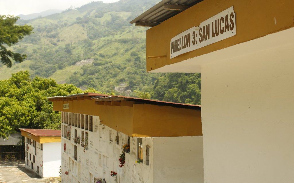 Vista exterior de un cementerio ubicado frente a una ladera montañosa cubierta de vegetación verde. En primer plano, la esquina superior de un bloque de nichos blanco con una franja ocre muestra el letrero "Pabellón 3: San Lucas". Atrás se observan otras estructuras similares con varias bóvedas.