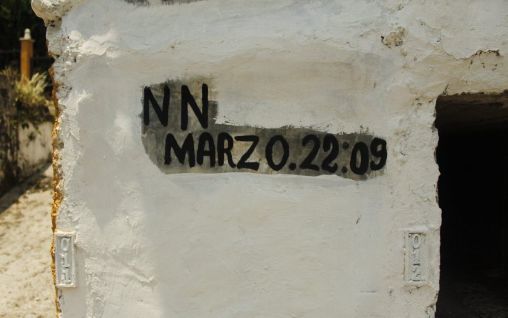 Primer plano del muro blanco y de textura rugosa de una bóveda de cementerio. Tiene pintada en letras negras la inscripción "NN MARZ O.22:09" sobre un fondo gris oscuro rectangular, indicando los restos de una persona no identificada. Justo al lado derecho se observa la abertura oscura de un nicho vacío.