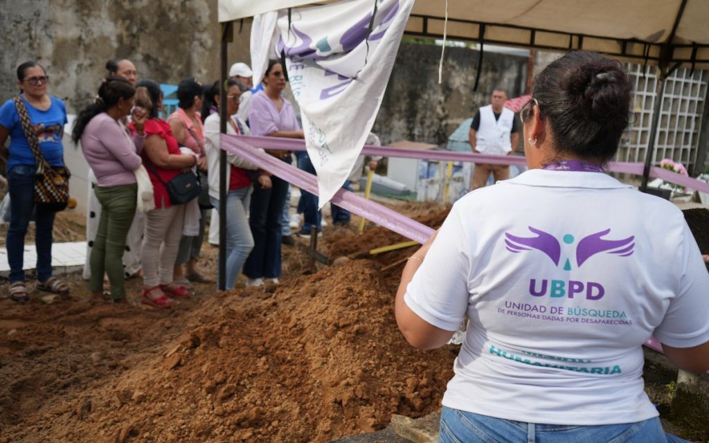 Fotografía en primer plano tomada desde atrás de una funcionaria de la Unidad de Búsqueda de Personas dadas por Desaparecidas (UBPD). Lleva una camiseta blanca con el logotipo azul y morado de la institución y el texto "UBPD - UNIDAD DE BÚSQUEDA DE PERSONAS DADAS POR DESAPARECIDAS / HUMANITARIA". Frente a ella se observa una fosa con montículos de tierra excavada y, al fondo, un grupo de familiares observando el procedimiento bajo la sombra de una carpa.