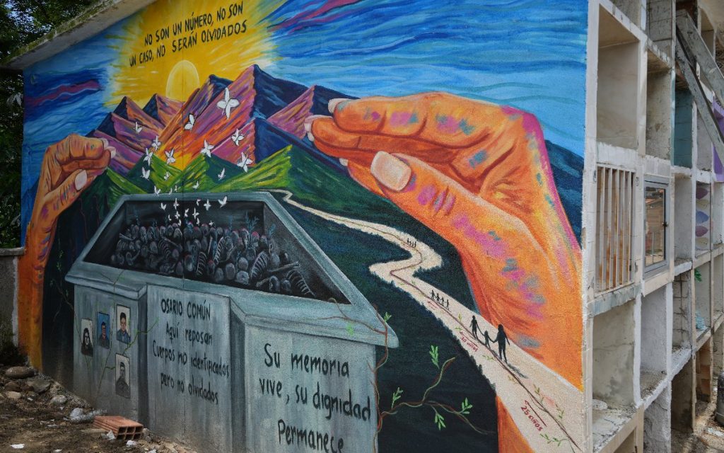 Fotografía de un mural pintado en un costado de la estructura de un cementerio. El mural ilustra dos grandes manos abiertas que protegen un osario común del cual emergen mariposas blancas que vuelan hacia un cielo azul con sol brillante. En la parte superior del mural se lee la frase: "No son un número, no son un caso, no serán olvidados". En el osario pintado aparece la frase "Osario común. Aquí reposan cuerpos no identificados pero no olvidados" y al lado "Su memoria vive, su dignidad permanece". A la derecha, un camino pintado muestra la silueta de familias caminando con las fechas de años transcurridos.