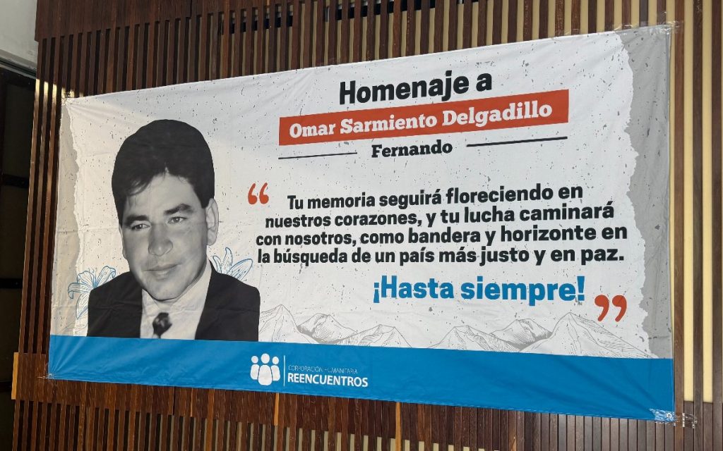 Pancarta de homenaje con el retrato en blanco y negro de un hombre. Contiene el texto: "Homenaje a Omar Sarmiento Delgadillo Fernando. Tu memoria seguirá floreciendo en nuestros corazones, y tu lucha caminará con nosotros, como bandera y horizonte en la búsqueda de un país más justo y en paz. ¡Hasta siempre!". En la parte inferior incluye el logotipo de la Corporación Humanitaria Reencuentros.
