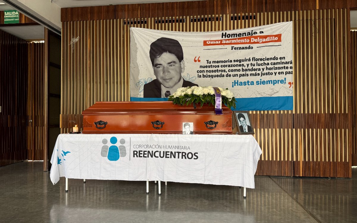 Ataúd de madera con un arreglo de flores blancas y una fotografía enmarcada de Omar Sarmiento en la parte superior. Reposa sobre una mesa cubierta con un mantel blanco que lleva el logotipo de la Corporación Humanitaria Reencuentros. De fondo, la pancarta del homenaje.