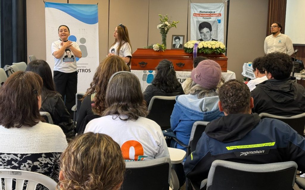 Una mujer se dirige al público asistente con las manos cruzadas sobre el pecho, vistiendo una camiseta estampada con el retrato de Omar Sarmiento. Detrás de ella se encuentra el ataúd y otras personas de pie acompañando la ceremonia.