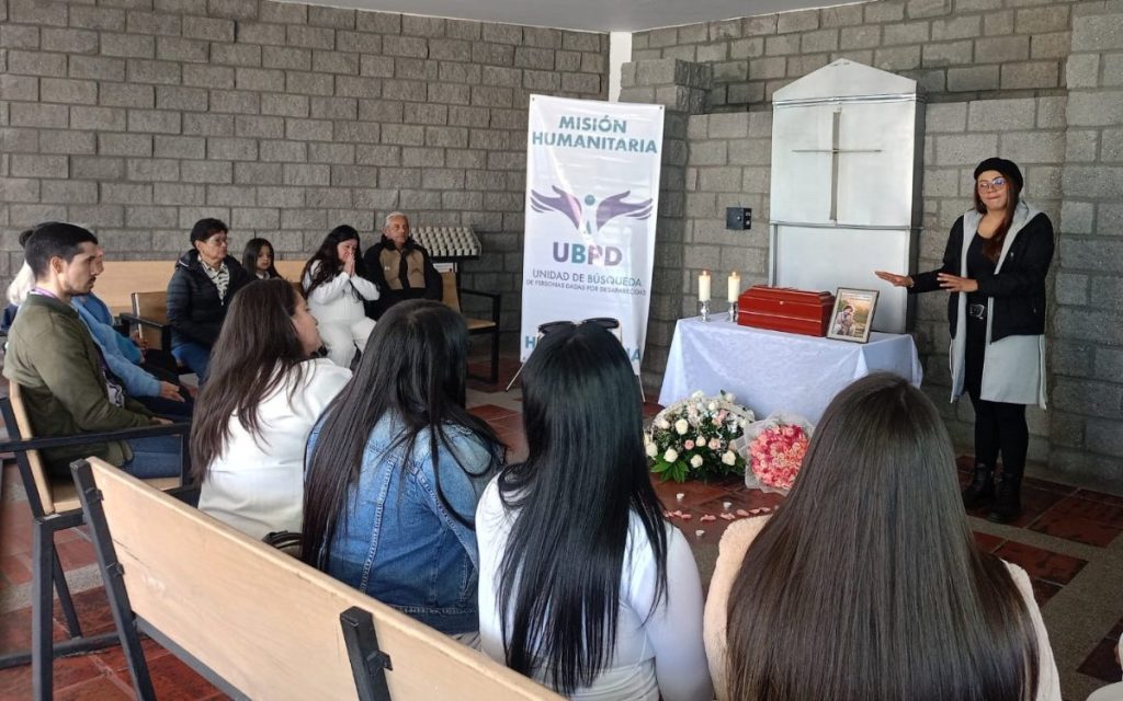 Una mujer de gafas y gorro negro de pie habla dirigiéndose a un grupo de personas sentadas en bancas de madera en un recinto religioso. Al fondo, sobre una mesa con mantel blanco, reposa un pequeño cofre funerario de madera de color caoba, una fotografía enmarcada, dos velas encendidas y un pendón de la UBPD con la frase "Misión Humanitaria". A los pies de la mesa hay arreglos florales.
