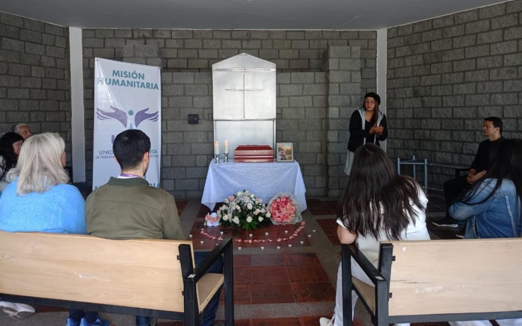 Vista panorámica desde la perspectiva de los asistentes sentados en bancas. En el centro, al fondo, un altar con mantel blanco sostiene un cofre funerario de madera, velas y una foto. En el suelo hay un arreglo floral y pétalos de rosas formando un camino. A la derecha, una mujer habla al grupo, mientras que a la izquierda destaca el pendón institucional de la UBPD "Misión Humanitaria".