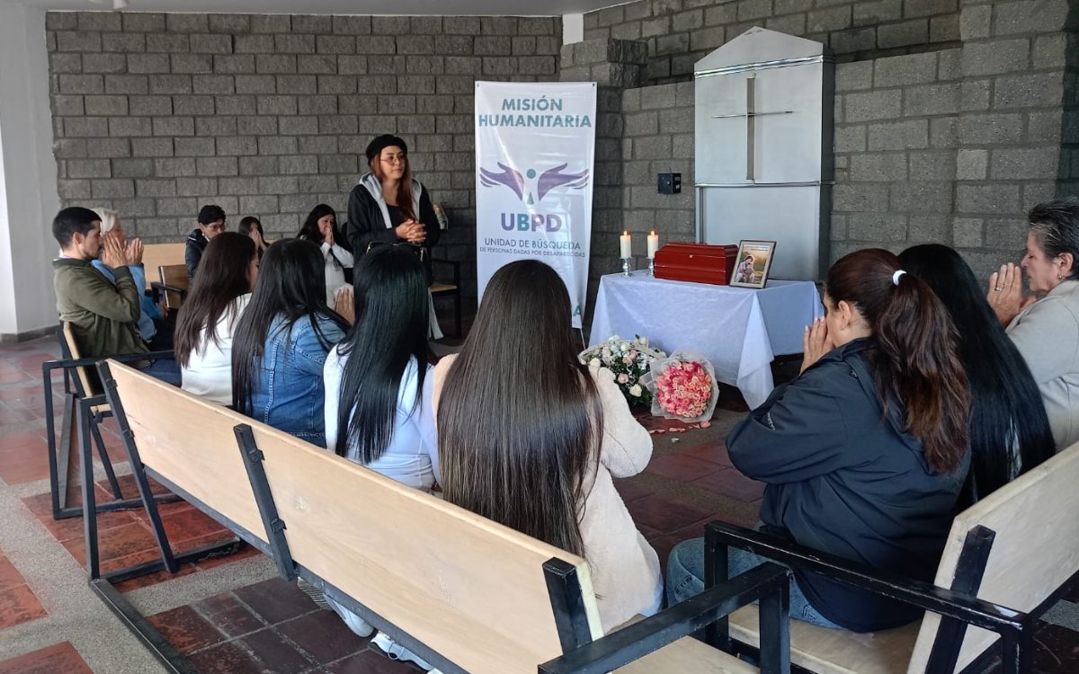 Plano lateral de los asistentes sentados en bancas de madera con las manos juntas en gesto de oración durante la entrega digna. Al fondo, la servidora de la UBPD permanece de pie junto al altar con el cofre funerario, la foto, las velas y el pendón de la misión humanitaria.