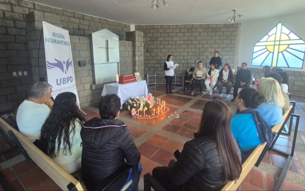 Vista amplia del recinto con paredes de bloque gris y un ventanal con vitral. Los asistentes y funcionarios están ubicados en semicírculo rodeando un camino de velas encendidas en el suelo de arcilla. Al fondo, una mujer lee un documento junto al altar con el cofre de madera y el pendón de la UBPD.