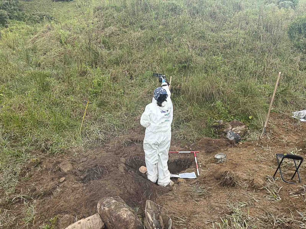 Vista superior de una profesional de la UBPD equipada con traje de bioseguridad blanco y guantes, tomando registro fotográfico de una fosa de excavación delimitada por una cuadrícula en terreno abierto.